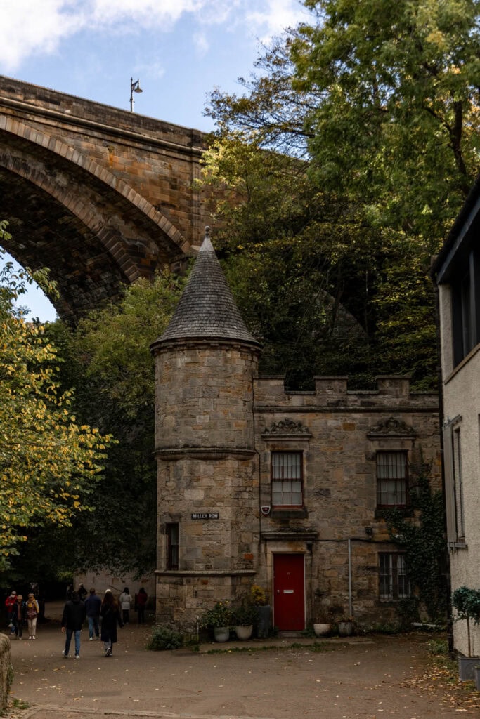 Charming historic stone building with turret under a bridge, surrounded by lush autumn foliage in a scenic travel setting.