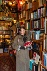 Vintage bookshop interior with shelves of colorful books and a smiling woman browsing, perfect for travel and literary destinations.