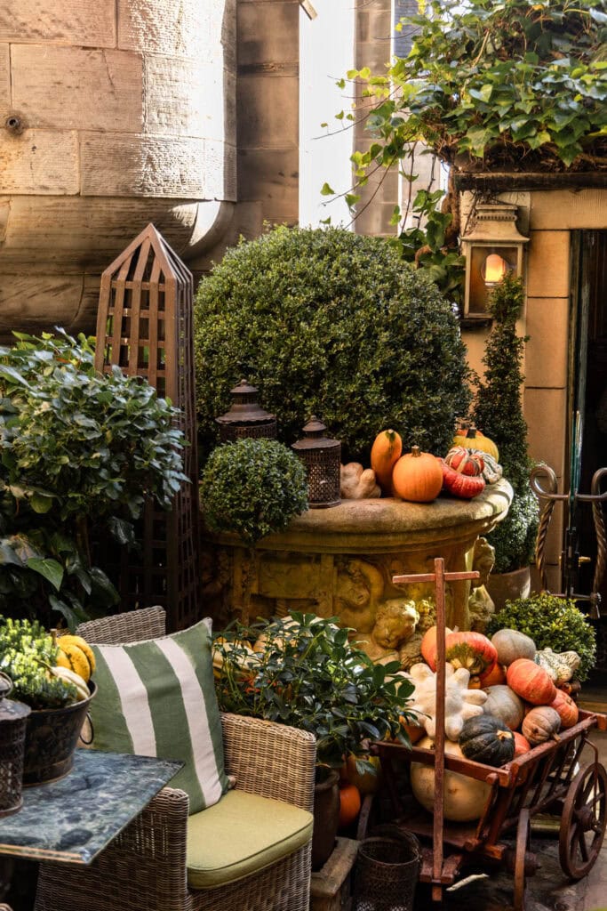 Pumpkin and fall decor on a stone fountain surrounded by lush greenery and vintage outdoor furniture.