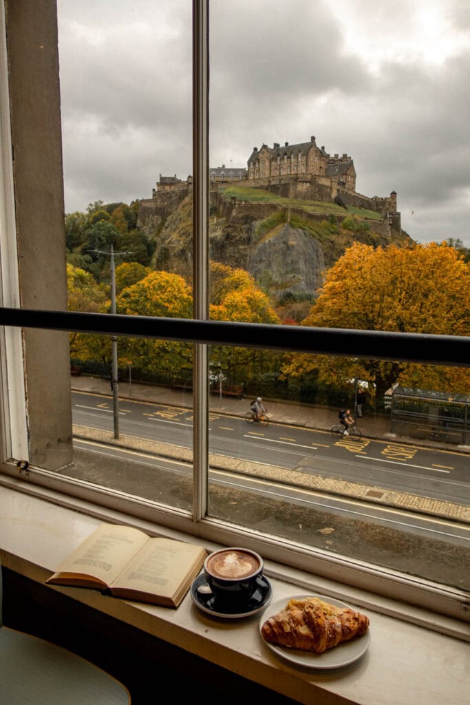 A scenic view of Edinburgh Castle from a cozy café window with a coffee and croissant on the sill.