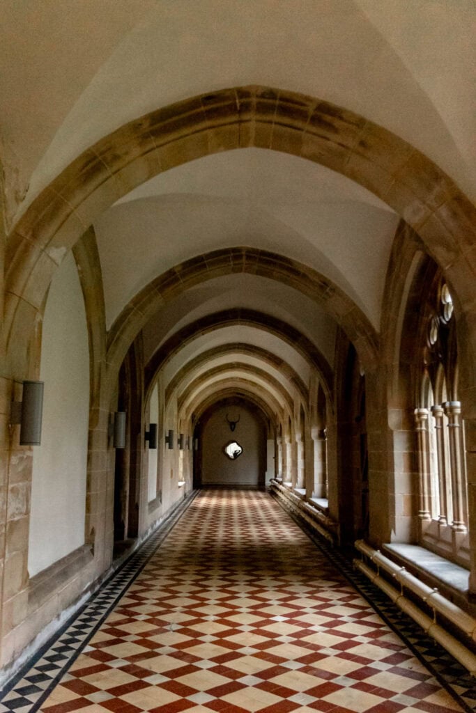 Stunning corridor with stone arches, gothic windows, and intricate tile flooring in a historic castle setting.
