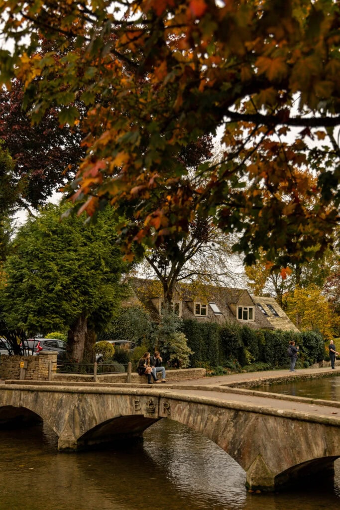 Colorful fall foliage over a quaint village with stone bridge, river, and residents relaxing outdoors.