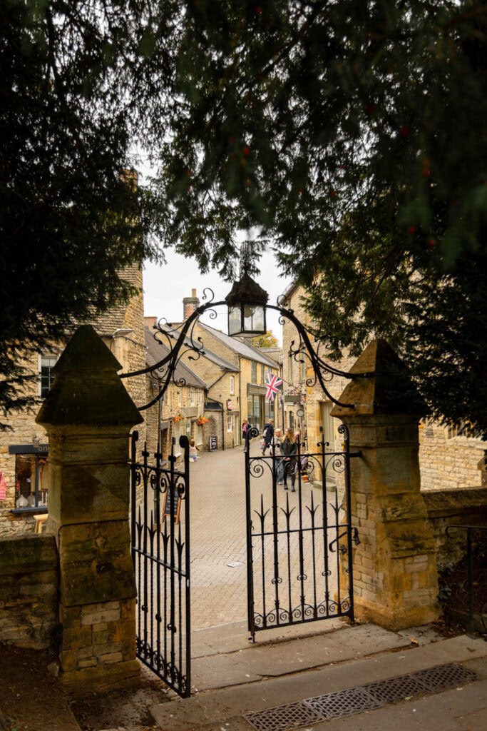 A picturesque view of a traditional English village street framed by an ornate gate and lush trees, perfect for travel enthusiasts.