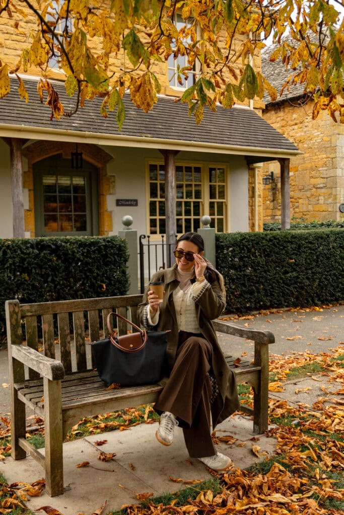 Scenic woman enjoying coffee outside a cozy cottage on autumn afternoon, surrounded by fallen leaves.