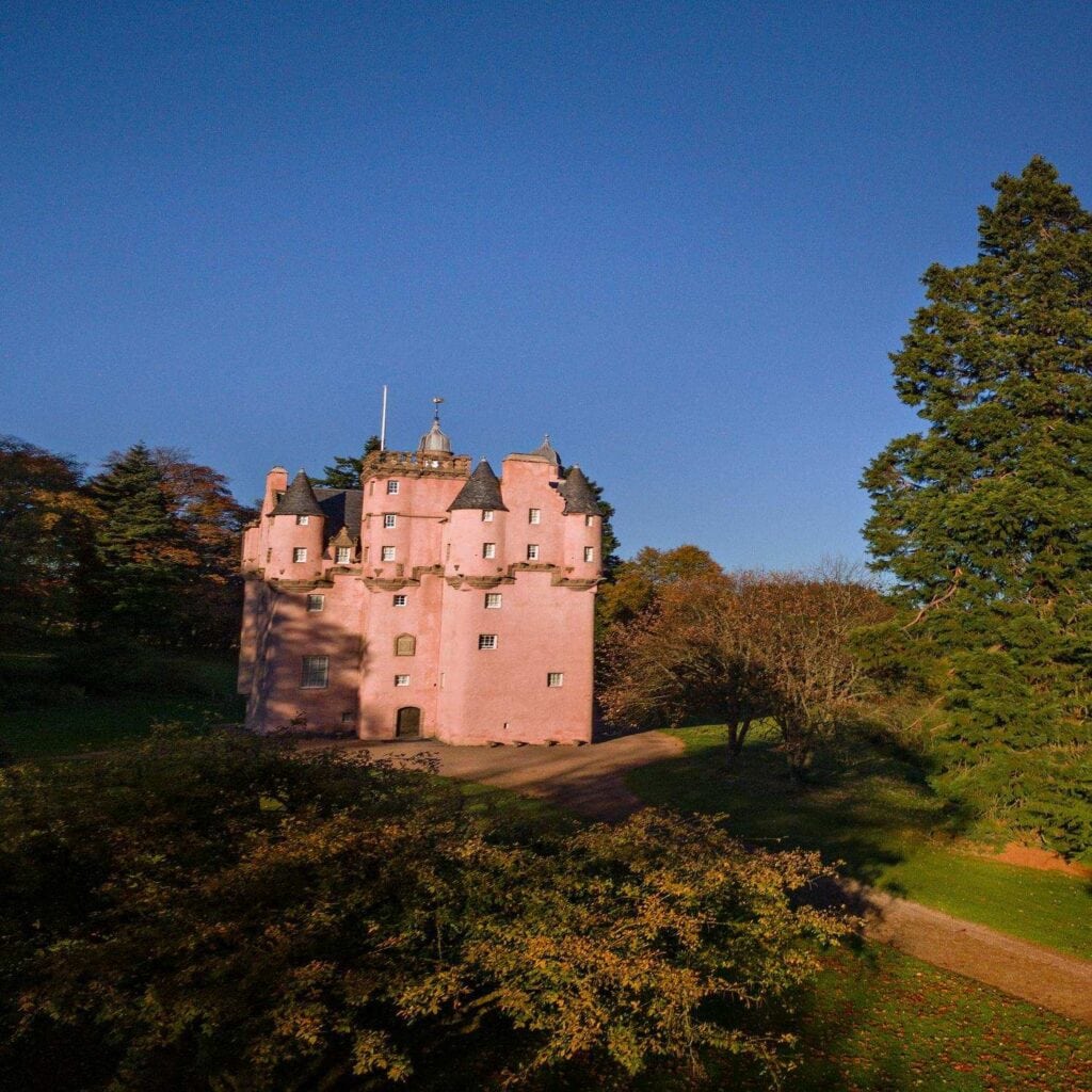 A tall, pink castle stands in the centre of the image, surrounded by lush trees and greenery. The building features turrets and small windows, with stone detailing around the roofline. The sky above is clear and blue, and the autumn foliage is visible in the foreground and background. The castle is set within landscaped grounds.