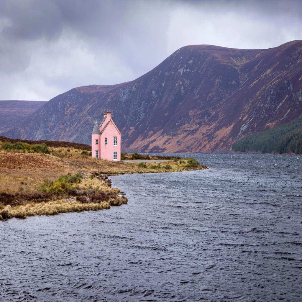 A charming pink house sits near the water's edge in the Scottish Highlands. The tranquil scene features a lake, rolling hills, and a cloudy sky, epitomising the serene, rugged beauty of Scotlands natural landscape.