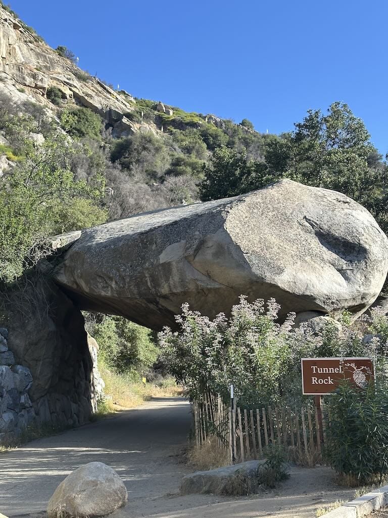 Large rock formation over a tunnel at Tunnel Rock, California, surrounded by greenery and a clear bl.