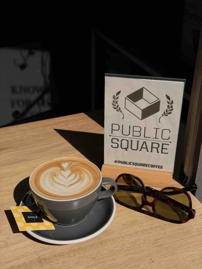 Coffee cup with latte art, sunglasses, and Public Square Coffee sign on wooden table.