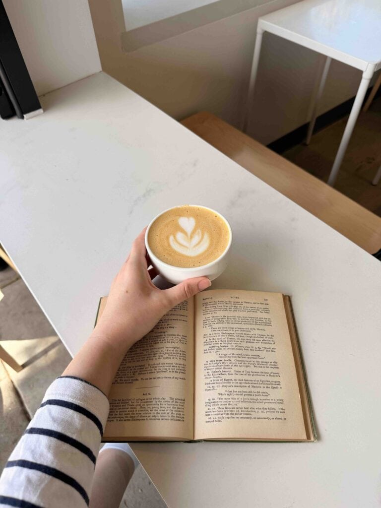 Person holding coffee with latte art and open book on white table.