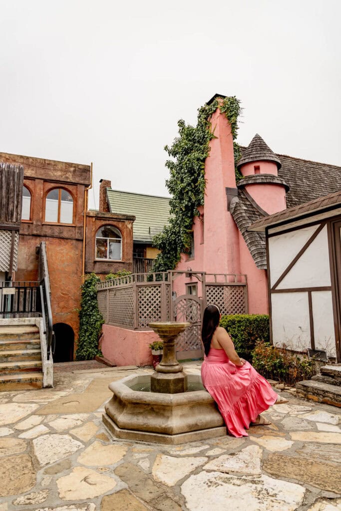 Charming woman in pink dress sitting by fountain in whimsical village setting.