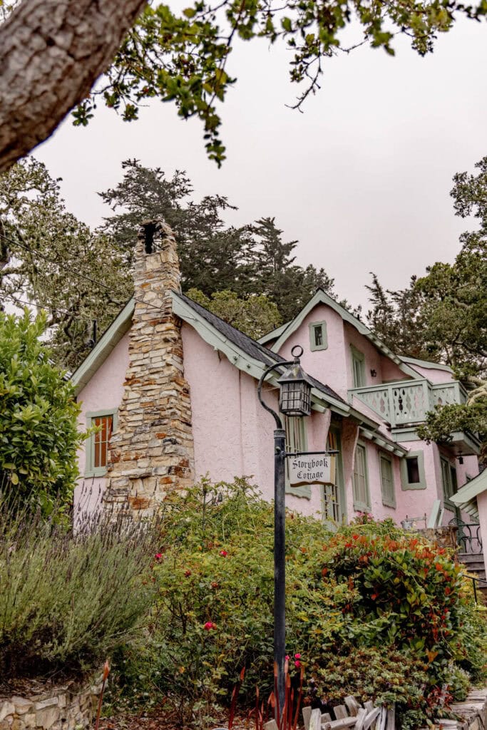 Pink cottage with stone chimney surrounded by lush greenery and flowering plants.