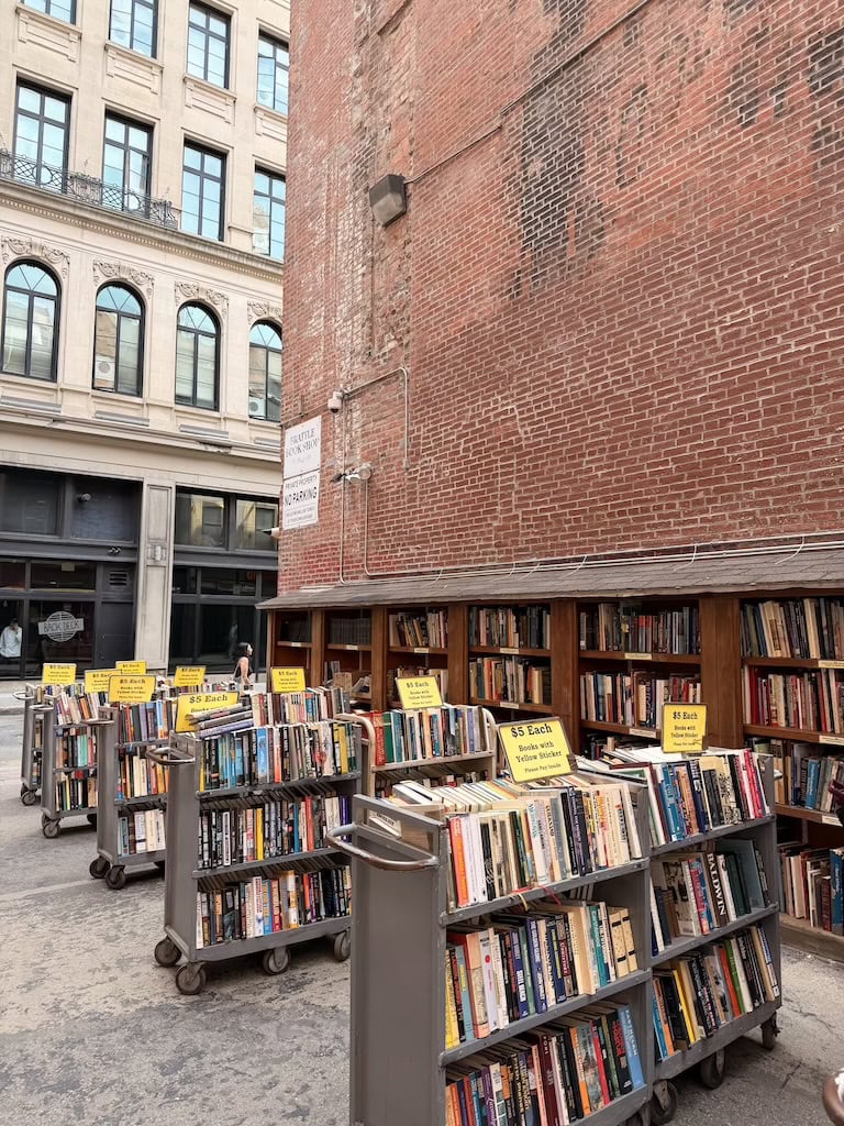 Street-side outdoor book sale with carts filled with books in front of a brick wall.