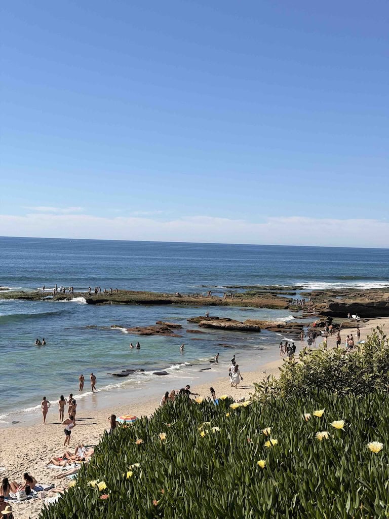 Scenic beach scene with people enjoying the sun, sand, and waves under a bright blue sky.